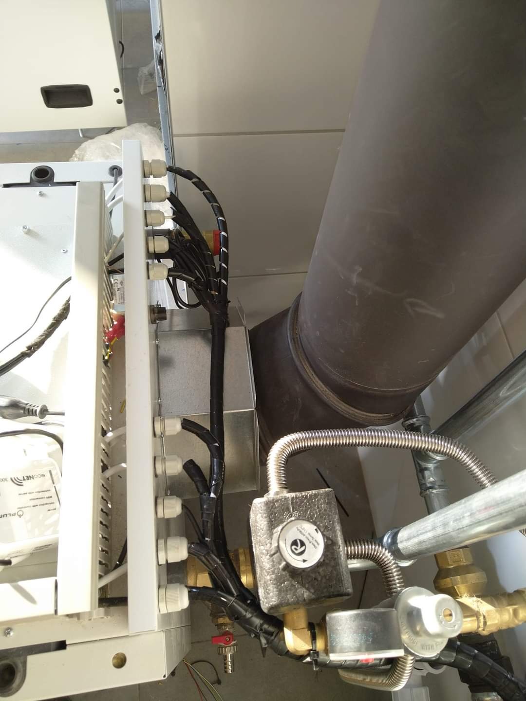 Fragment instalacji grzewczej z widocznym kotłem, rurami, zaworami i przewodami elektrycznymi w trakcie podłączenia. Widoczna izolacja termiczna na zaworze.