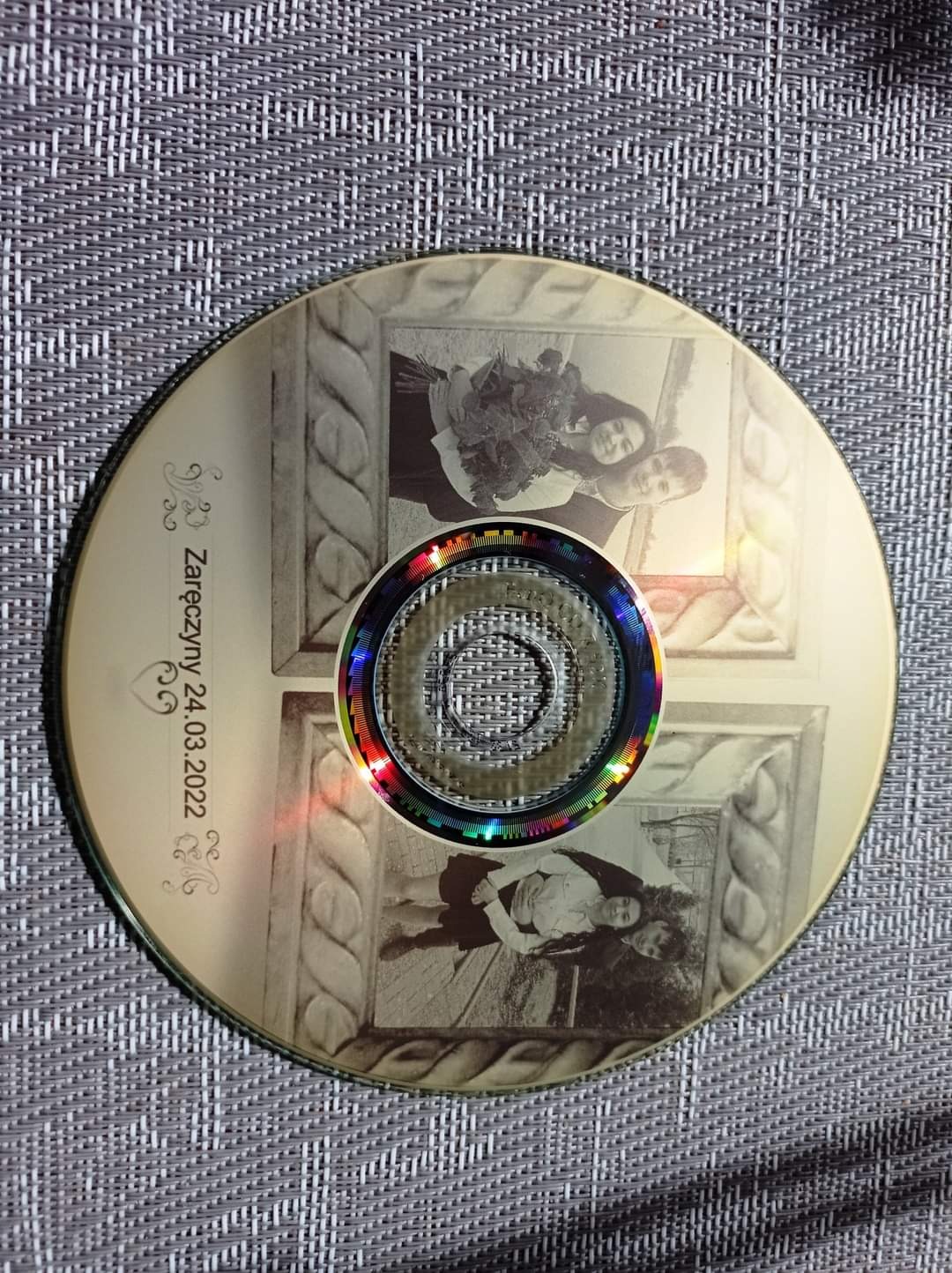 Płyta CD z nadrukowanymi czarno-białymi zdjęciami pary w ozdobnych ramkach i datą zaręczyn, leżąca na teksturowanej powierzchni.