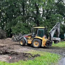 Żółta koparko-ładowarka Volvo BL71 na placu budowy, z widocznym numerem telefonu na kabinie, wykonująca prace ziemne na tle zieleni.