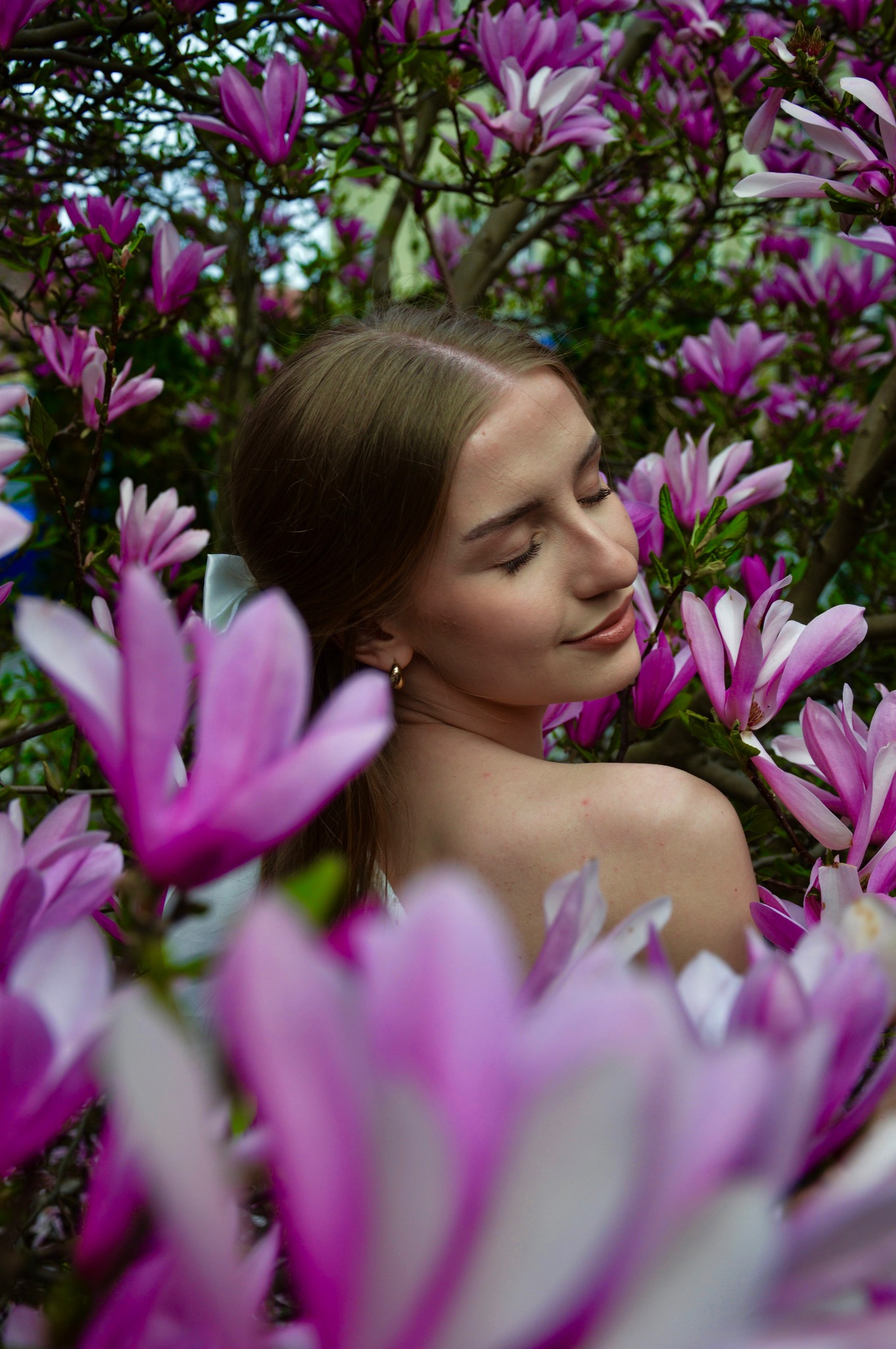 Portret kobiety z zamkniętymi oczami, otoczonej kwitnącymi magnoliami w odcieniach różu i fioletu. Włosy upięte białą kokardą, na uchu złoty kolczyk.