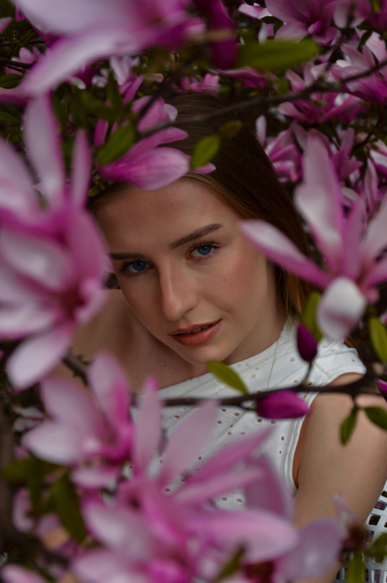 Portret młodej kobiety o niebieskich oczach, otoczonej kwitnącymi różowymi magnoliami, w białej ażurowej bluzce.