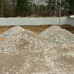 Dwa pryzmy gruzu z resztkami cegieł na placu budowy, w tle betonowe ogrodzenie panelowe i drzewa.