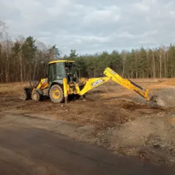 Żółta koparko-ładowarka JCB podczas prac ziemnych na terenie z widocznymi śladami opon i lasem w tle.