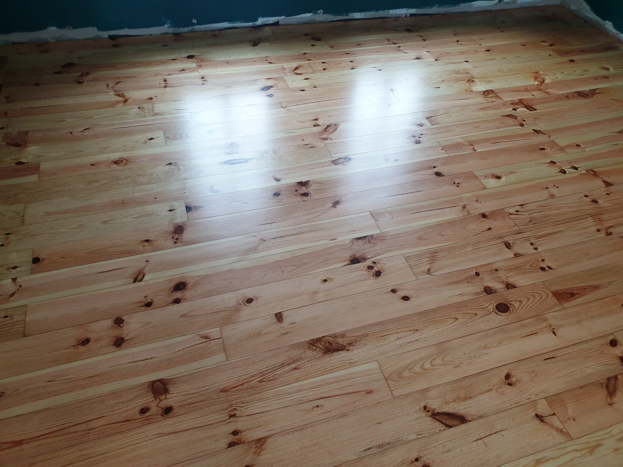 Jasny, wycyklinowany parkiet sosnowy z widocznymi słojami i sękami, świeżo położony na podłodze w trakcie remontu, z widocznymi śladami malowania na ścianie.