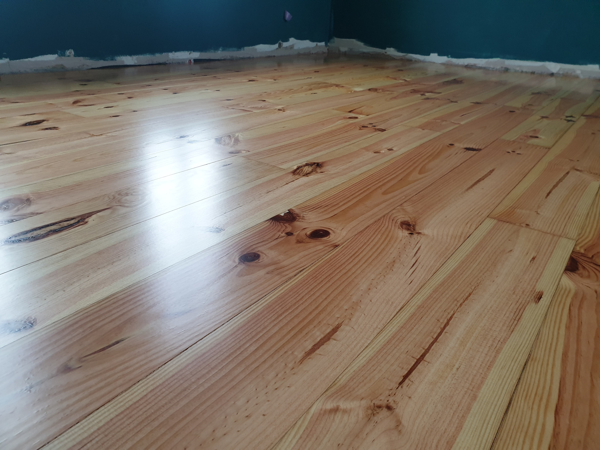 Świeżo polakierowany parkiet z widocznymi słojami drewna i odblaskiem światła, ściana pomalowana na ciemny kolor z widocznymi śladami po malowaniu na listwie przypodłogowej.