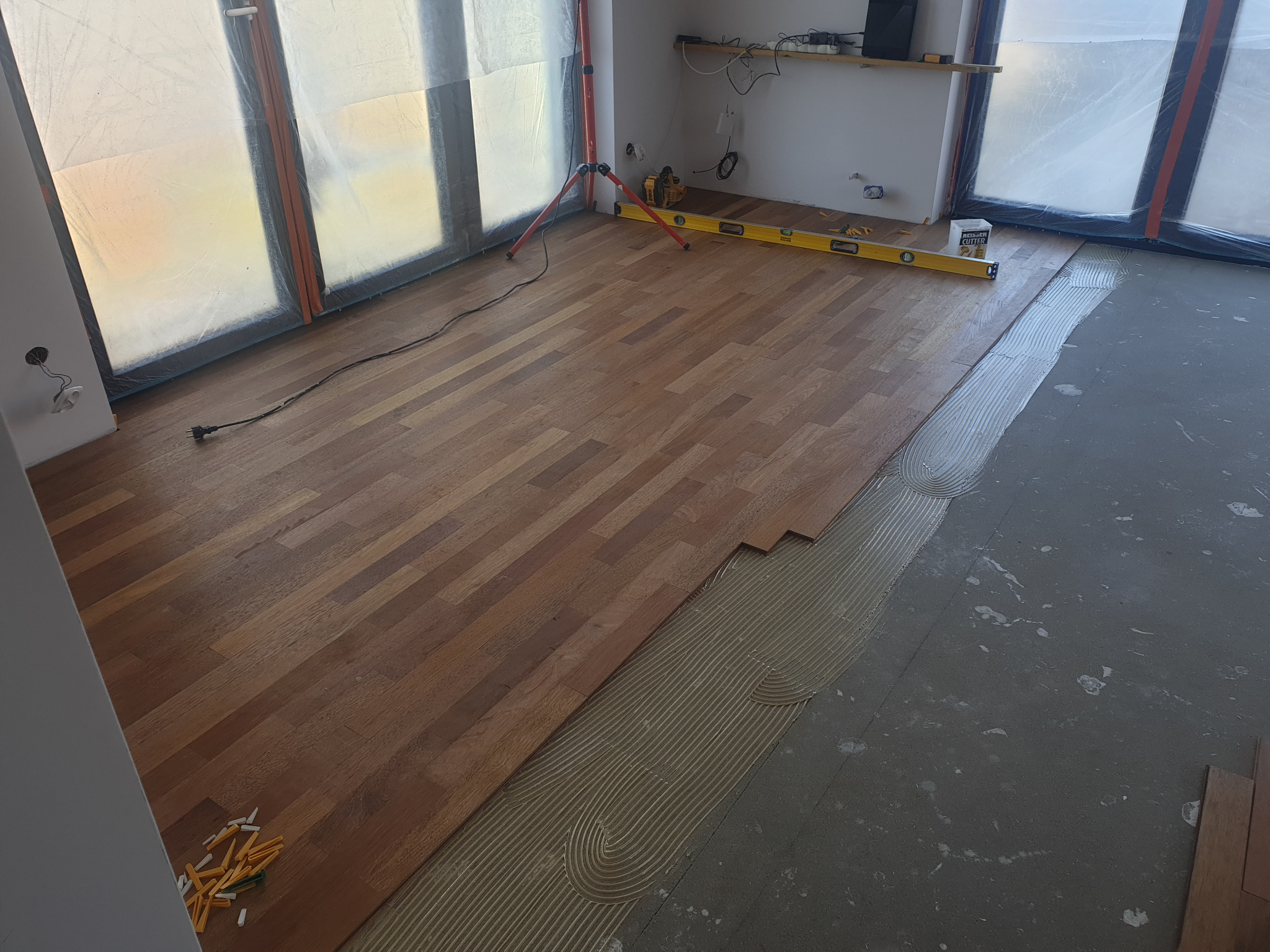 Układanie parkietu z egzotycznego drewna; widoczny klej rozprowadzony na podłożu, poziomica, kliny dystansowe i zabezpieczone folią okna.