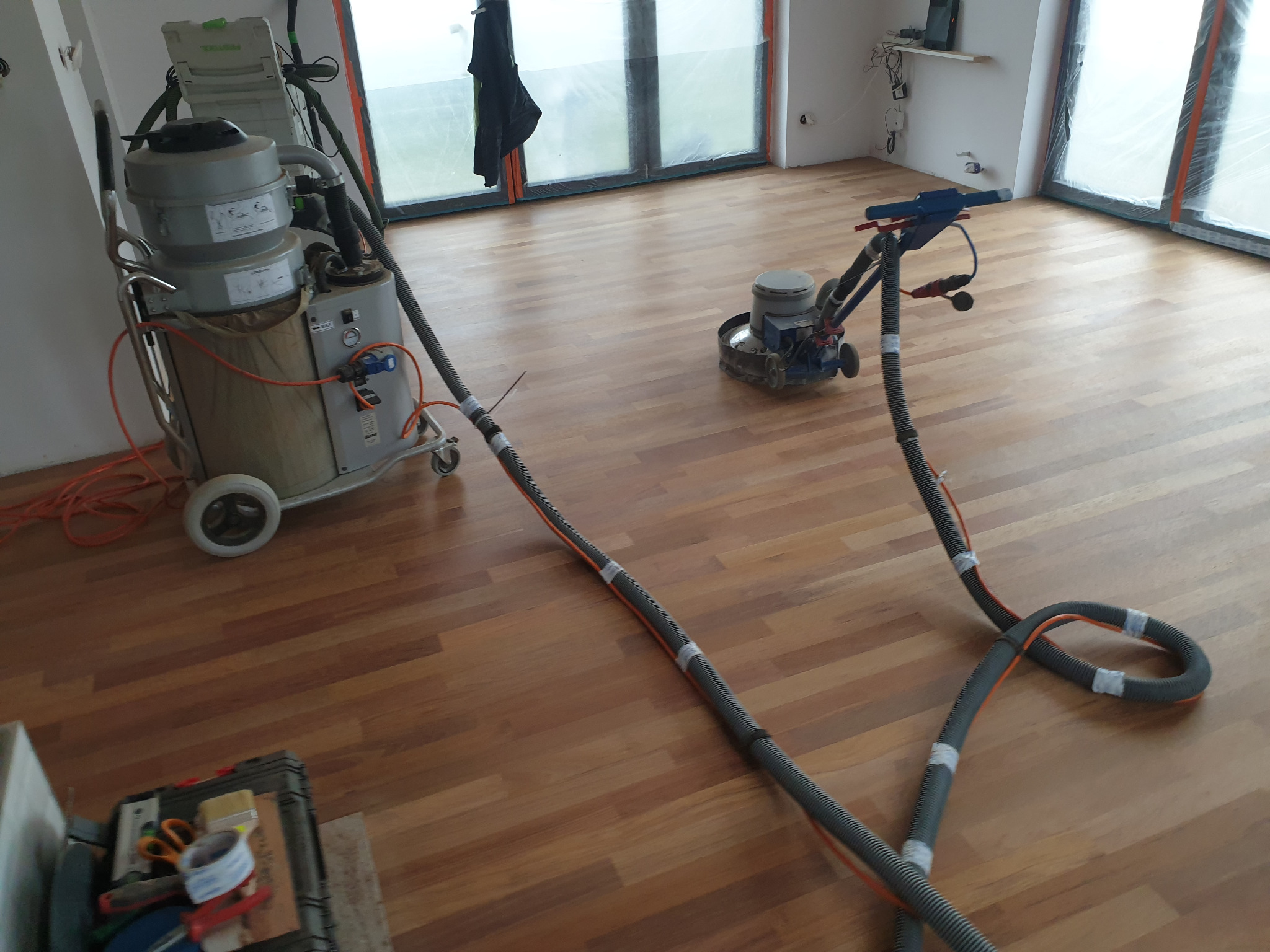 Parkiet z drewna egzotycznego po obróbce szlifierskiej, widoczne maszyny do cyklinowania parkietu i odkurzacz przemysłowy z podłączonymi wężami.