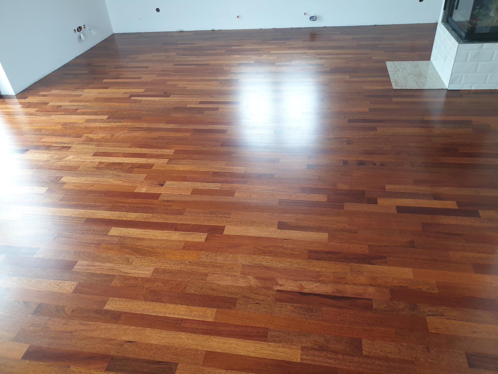 Błyszcząca, nowo położona drewniana podłoga z parkietu w odcieniach brązu, ułożona w cegiełkę, z widocznym fragmentem kominka z białych płytek w tle.