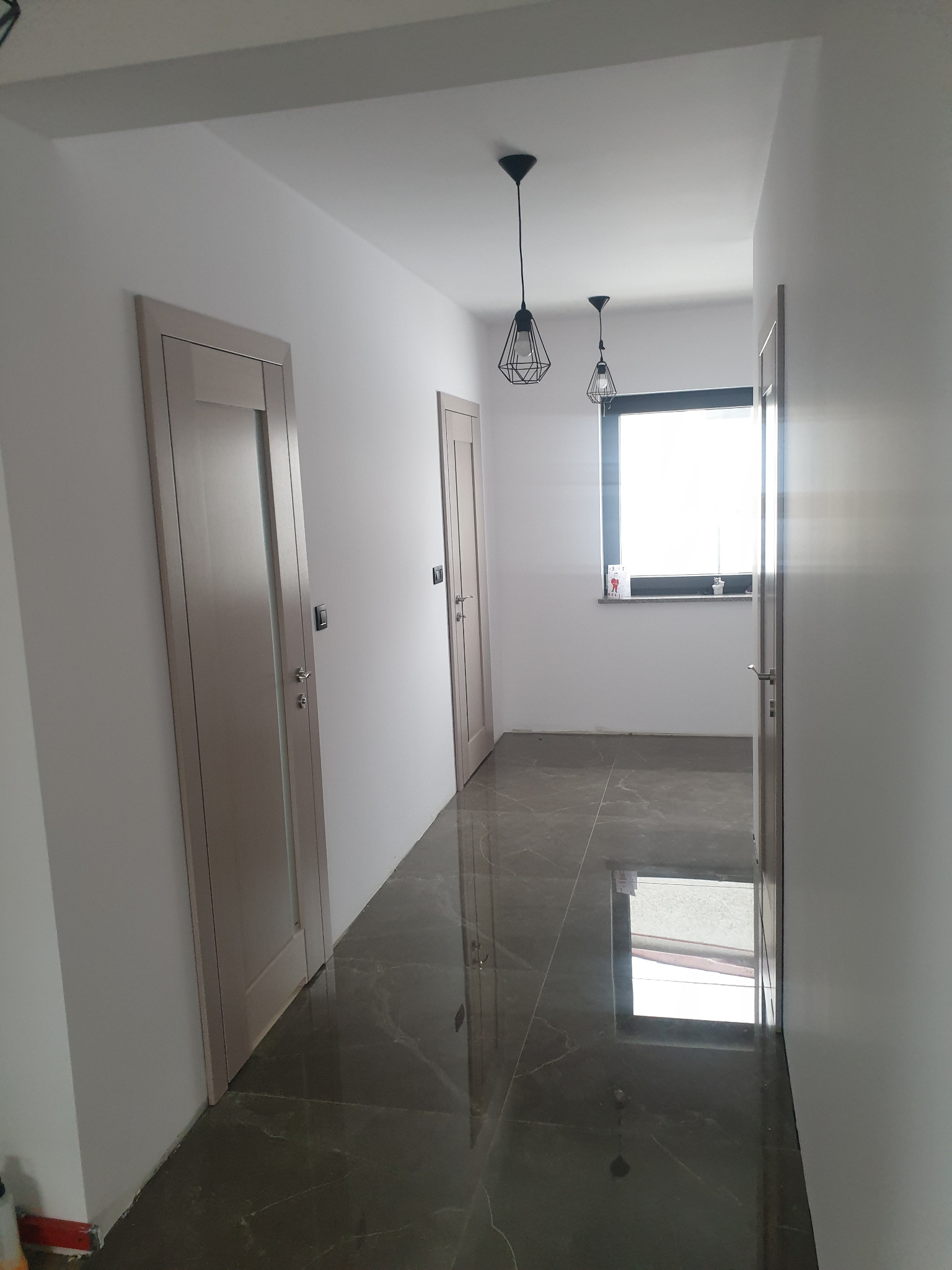 Korytarz po remoncie z szarą marmurową podłogą, trzema beżowymi drzwiami z srebrnymi klamkami i nowoczesnymi lampami wiszącymi w kształcie klatek.