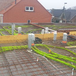 Investa - Zbrojenie płyty fundamentowej z zielonymi rurami instalacji ogrzewania podłogowego, widoczne pionowe rury kanalizacyjne zabezpieczone folią, fragmenty szalunków z żółtej płyty i sąsiednie budynki...