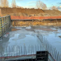 Zalany betonem fundament z wystającymi prętami zbrojeniowymi i żółtymi szalunkami, widok na wykop z pomarańczową siatką zabezpieczającą i hałdą ziemi w tle.