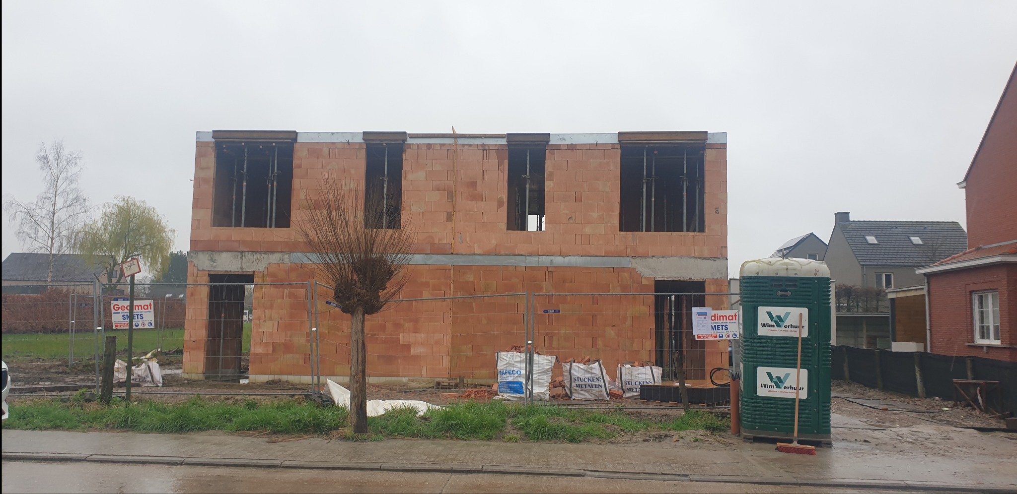 Budowa domu murowanego w trakcie wznoszenia ścian z pustaków ceramicznych, widoczne worki z zaprawą, zabezpieczenia budowy i przenośna toaleta.