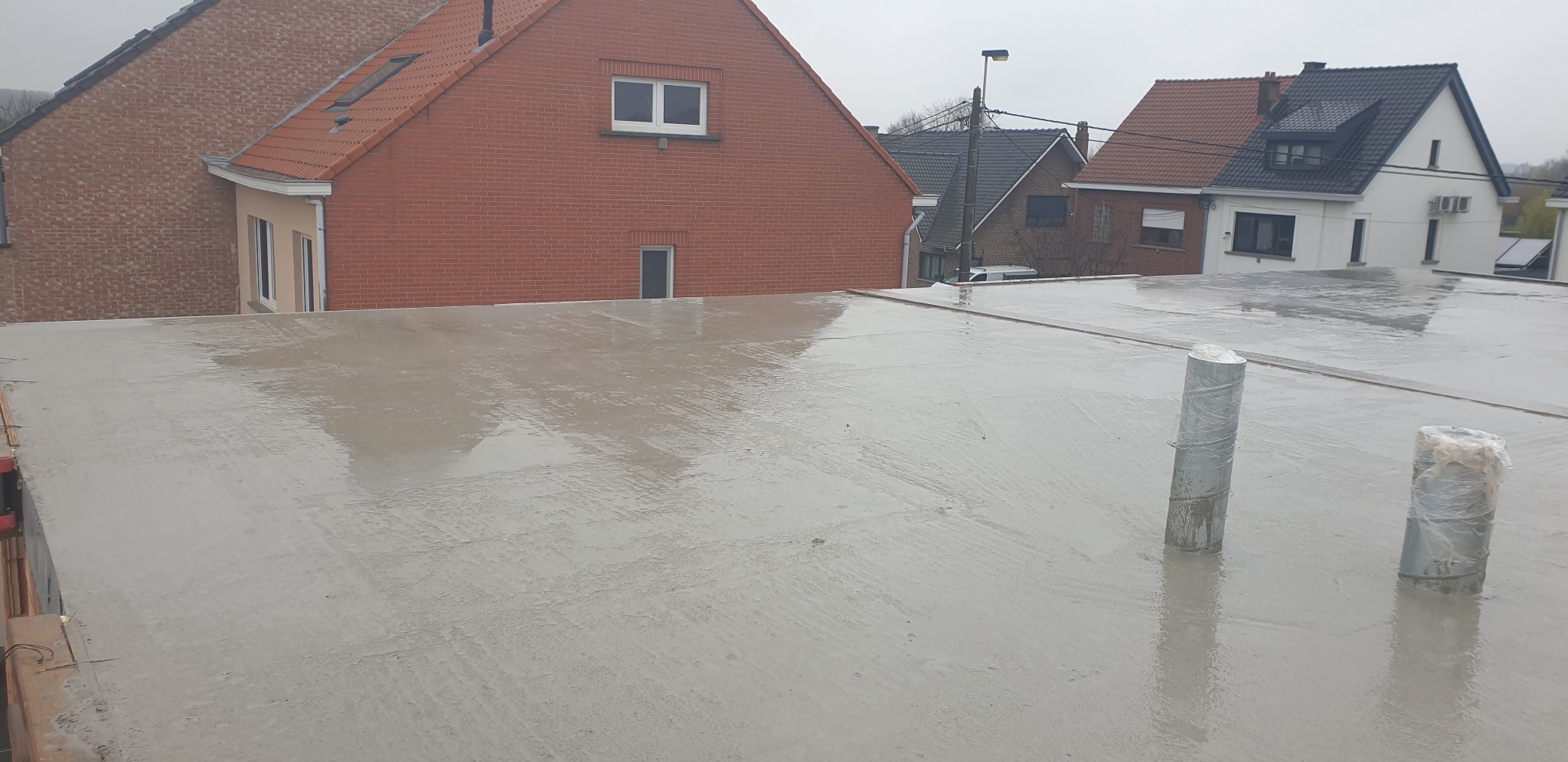 Świeżo wylany, mokry strop betonowy na budowie domu jednorodzinnego, widoczne rury instalacyjne zabezpieczone folią, w tle zabudowa mieszkalna.