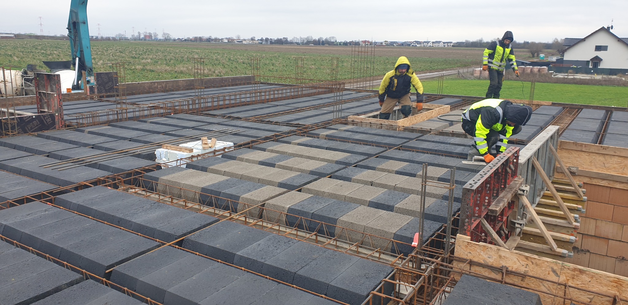 Budowa stropu Teriva: robotnicy w odblaskowych kamizelkach układają pustaki pomiędzy żebrami konstrukcji, widoczne szalunki i zbrojenie, w tle krajobraz wiejski.