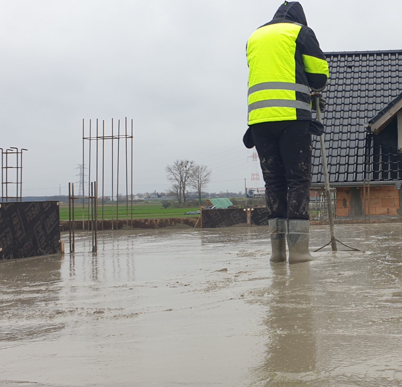 Pracownik w odblaskowej kamizelce i gumowcach wyrównuje świeżo wylany beton na fundamencie domu, widoczne zbrojenia i deski szalunkowe, w tle zarys domu z czarną dachówką.