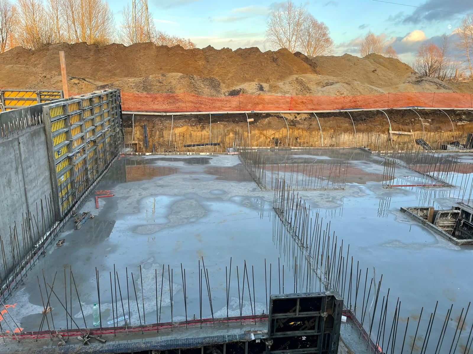 Zalany betonem fundament z wystającymi prętami zbrojeniowymi i żółtymi szalunkami, widok na wykop z pomarańczową siatką zabezpieczającą i hałdą ziemi w tle.