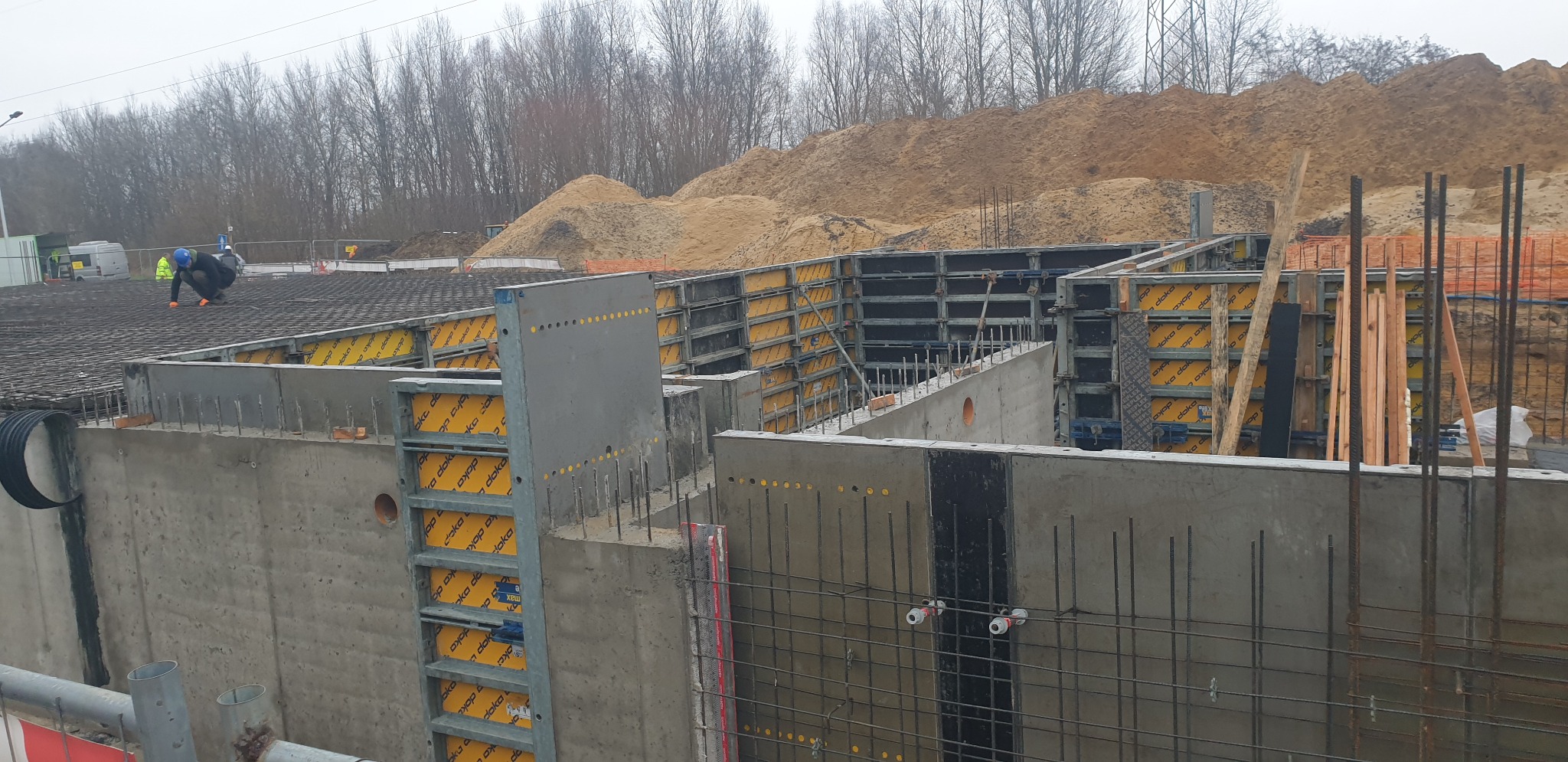 Wykop pod budynek z szalunkami, zbrojeniami i pracownikiem w kasku układającym zbrojenie na górze. W tle hałda piasku i drzewa.