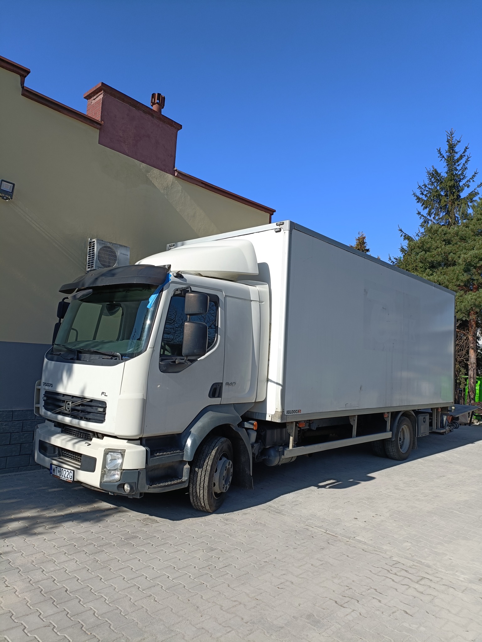 Biała ciężarówka Volvo FL z zabudową kontenerową na brukowanym placu, widoczny klimatyzator na ścianie budynku w tle, słoneczny dzień.