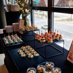 ATEITIS SPÓŁKA Z OGRANICZONĄ ODPOWIEDZIALNOŚCIĄ - Stół cateringowy z różnorodnymi przekąskami: mini tartaletki z pomidorami, kanapeczki z pastą, sałatki w słoiczkach i talerze z deserami, ustawiony przy oknie z widokiem na ulicę.