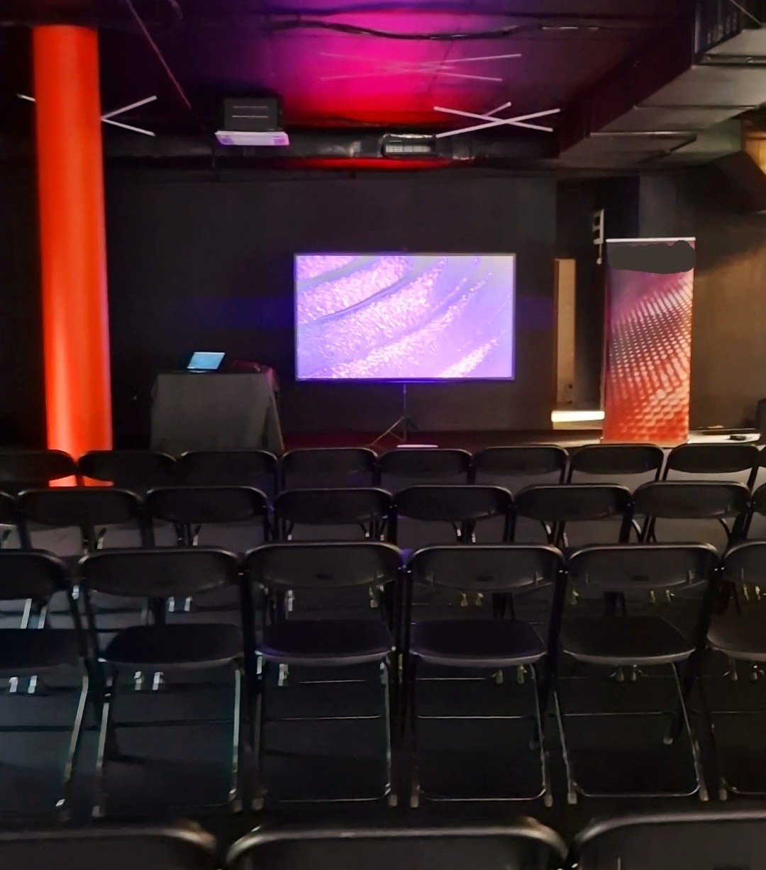 Sala konferencyjna z rzędami czarnych krzeseł, ekranem projekcyjnym wyświetlającym abstrakcyjny obraz w odcieniach fioletu, czerwona kolumna po lewej stronie, oświetlenie sceniczne w kolorze...