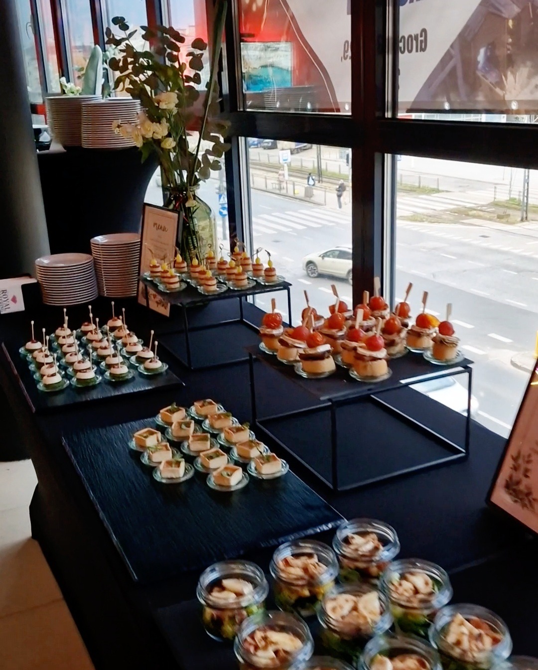Stół cateringowy z różnorodnymi przekąskami: mini tartaletki z pomidorami, kanapeczki z pastą, sałatki w słoiczkach i talerze z deserami, ustawiony przy oknie z widokiem na ulicę.