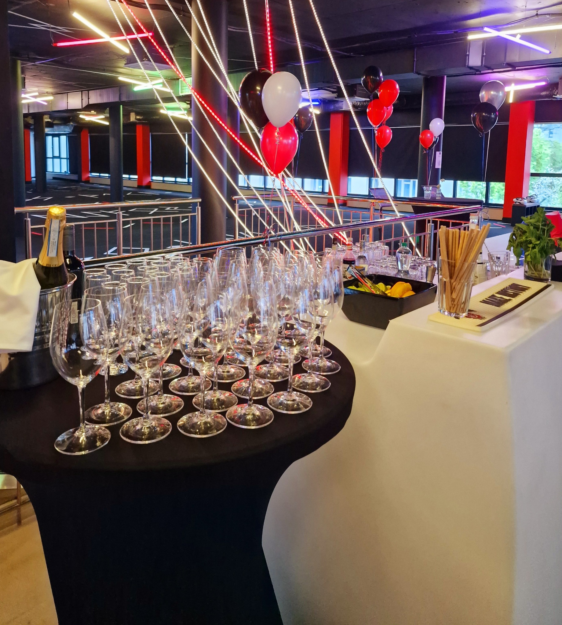 Przygotowany bar z kieliszkami, szampanem w wiaderku z lodem, przekąskami i słomkami na tle dekoracji świetlnych i balonów w sali.