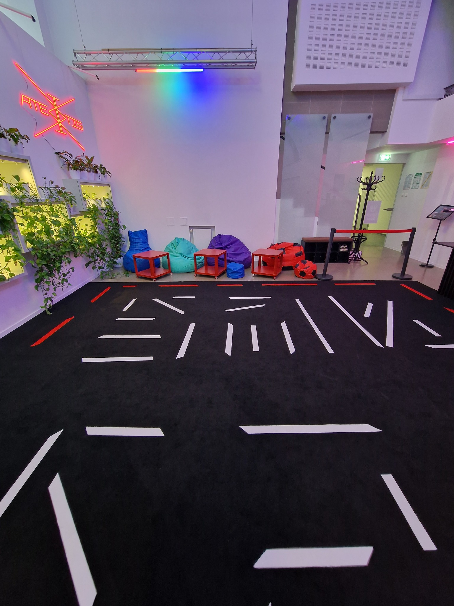 Przestrzeń eventowa z czarną wykładziną, białymi liniami tworzącymi wzór na podłodze, kolorowymi pufami do siedzenia, neonem na ścianie i roślinami w doniczkach