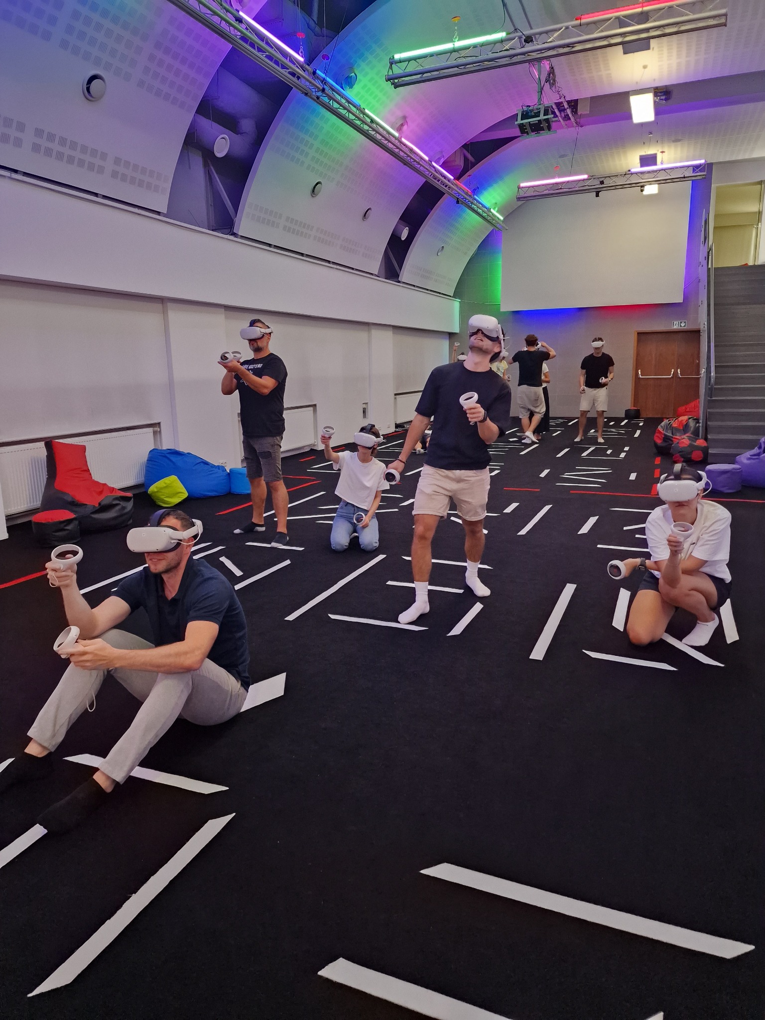 Grupa osób w goglach VR, trzymających kontrolery, uczestniczy w sesji wirtualnej rzeczywistości w przestronnym pomieszczeniu z kolorowym oświetleniem sufitowym i geometrycznym wzorem na podłodze.