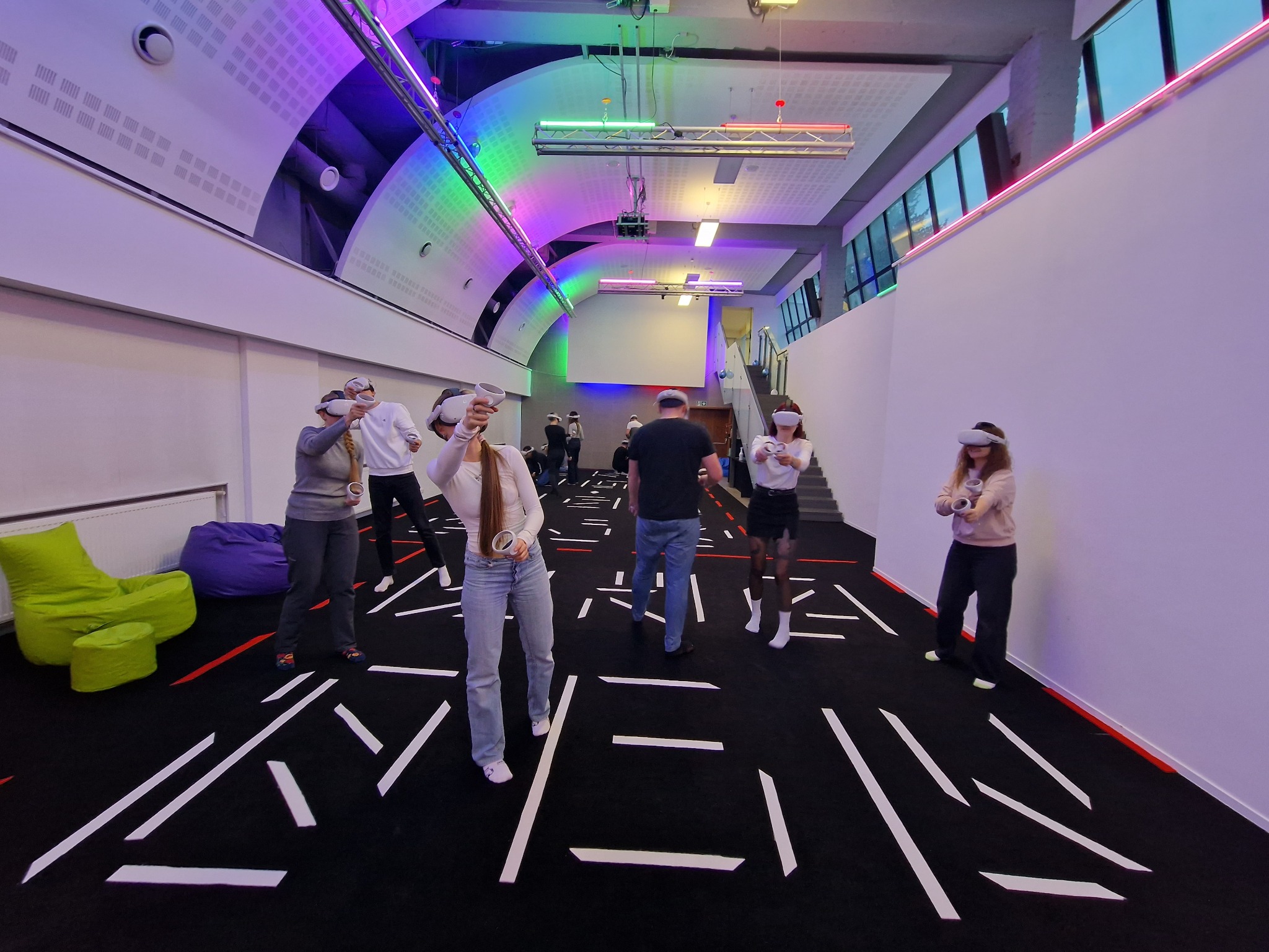 Grupa osób w goglach VR, trzymających kontrolery, w przestronnym pomieszczeniu z oświetleniem LED i geometrycznym wzorem na podłodze.
