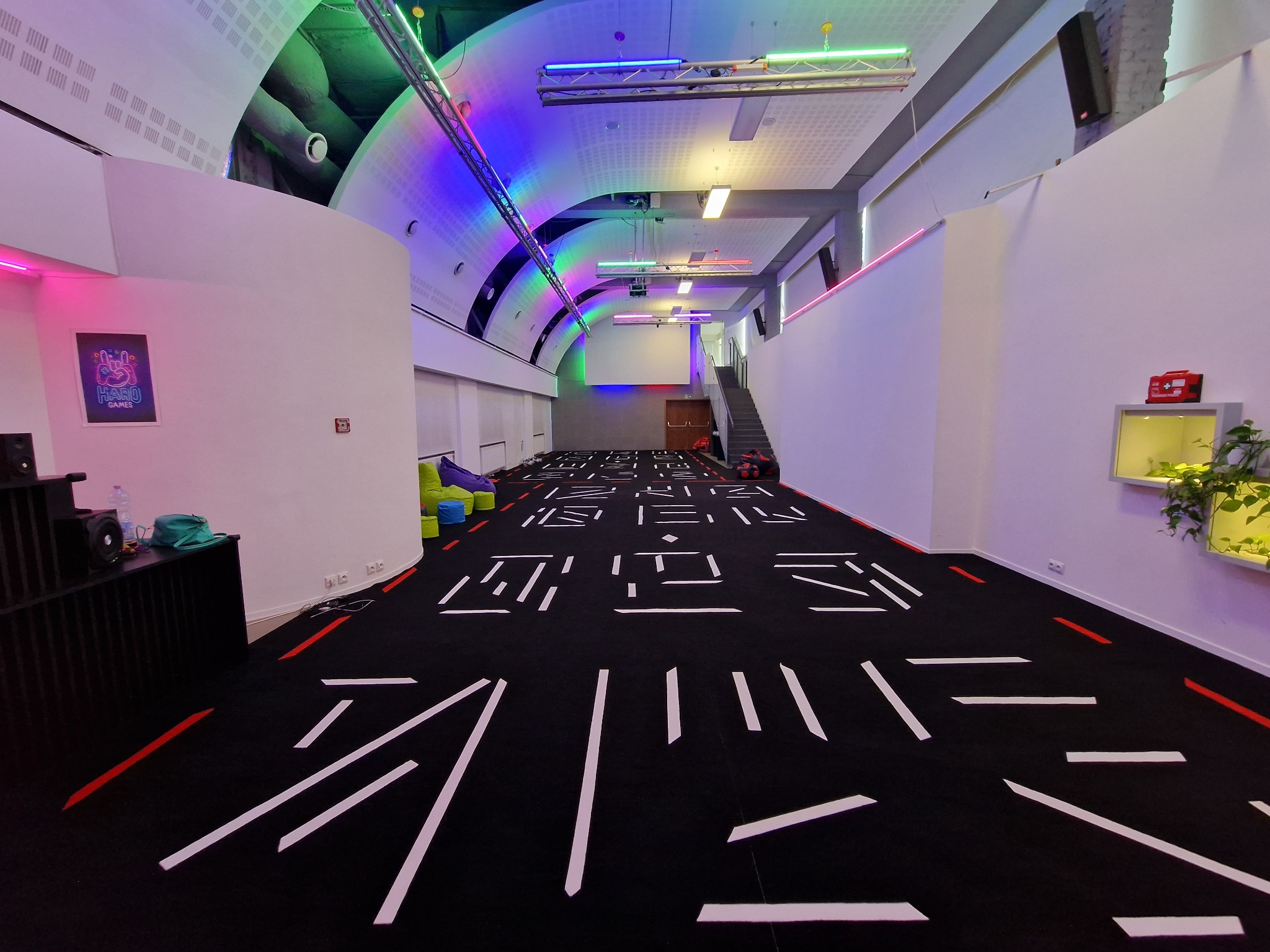 Przestronne wnętrze sali z czarną podłogą ozdobioną białymi i czerwonymi liniami, kolorowe oświetlenie sufitowe, beanbagi w tle, oraz plakat z logo 'HARU GAMES' na ścianie.