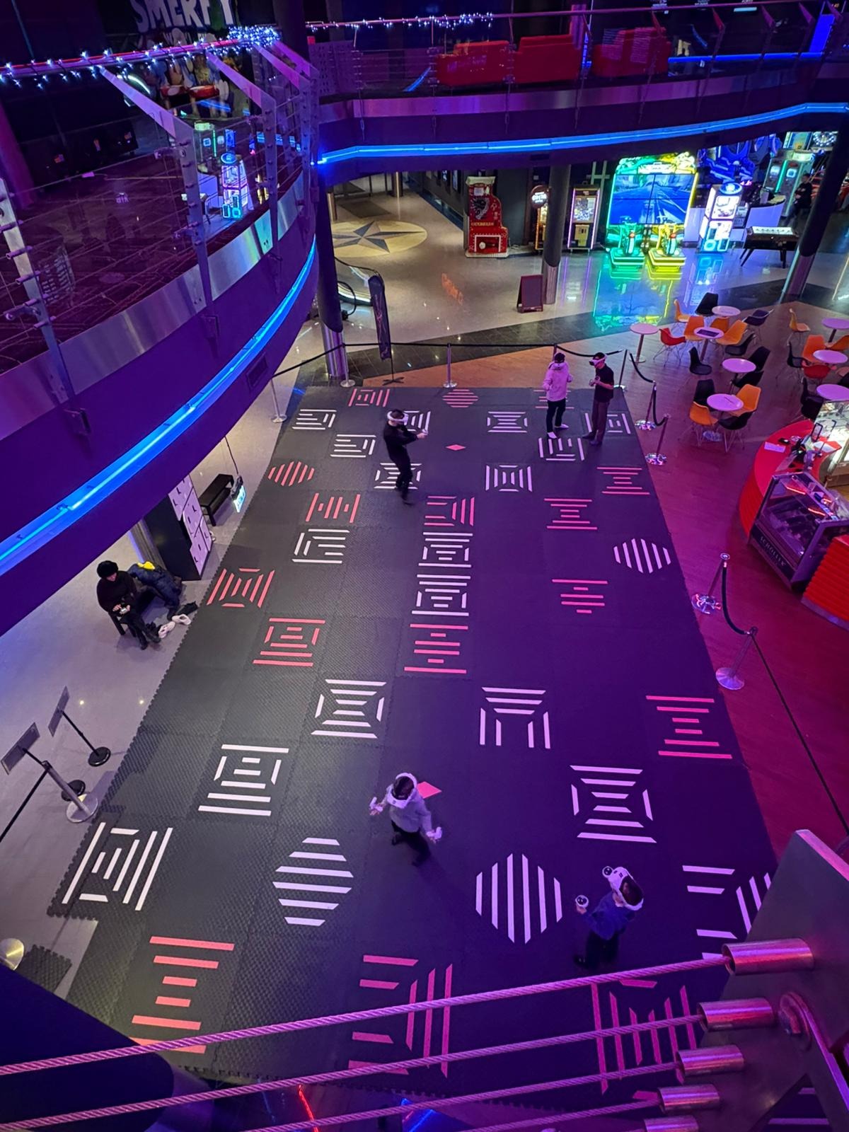 Wnętrze centrum rozrywki z osobami w goglach VR na interaktywnej podłodze. Widok z góry, oświetlenie neonowe, automaty do gier w tle.