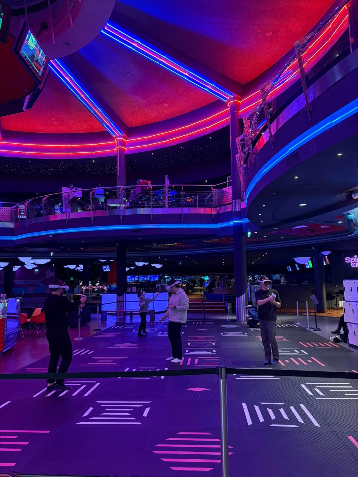 Wnętrze centrum rozrywki z neonowym oświetleniem, gdzie osoby w goglach VR uczestniczą w grze. Widoczne elementy scenografii i wyposażenia.