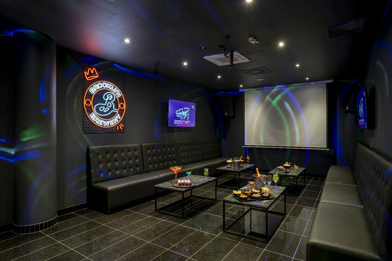 Nowoczesna sala imprezowa w ciemnej tonacji z neonem Brooklyn Brewery, skórzanymi kanapami i stolikami z przekąskami, oświetlona kolorowymi światłami.