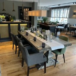 Meg@wat Jacek Remplakowski - Wnętrze restauracji z drewnianym stołem nakrytym do uroczystego obiadu, oświetlonym przez podłużną lampę wiszącą i lampy punktowe na suficie.