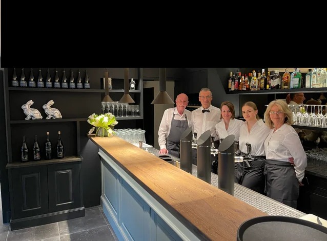 Zespół barmanów i kelnerów w eleganckim lokalu z czarnymi elementami wystroju, widoczne dystrybutory do piwa i półki z alkoholem.