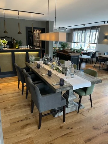 Wnętrze restauracji z drewnianym stołem nakrytym do uroczystego obiadu, oświetlonym przez podłużną lampę wiszącą i lampy punktowe na suficie.