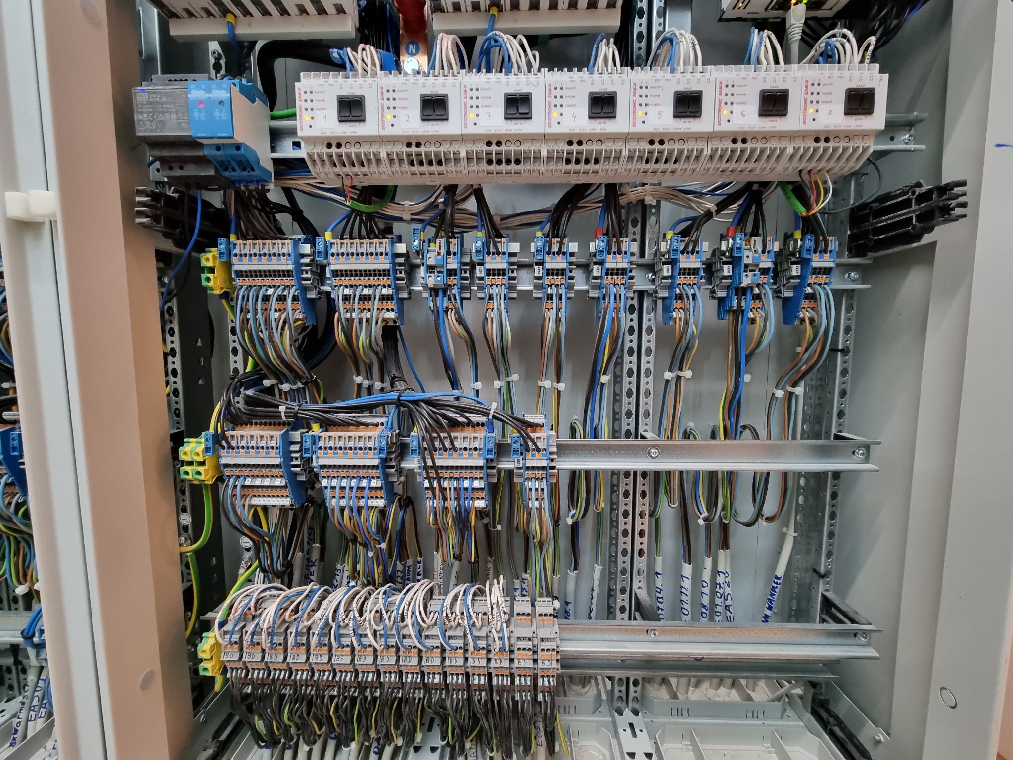 Szafa sterownicza z widocznymi elementami elektrycznymi, w tym listwy zaciskowe, przewody w różnych kolorach oraz moduły sterujące z diodami LED i przełącznikami, zamontowana w metalowej obudowie.