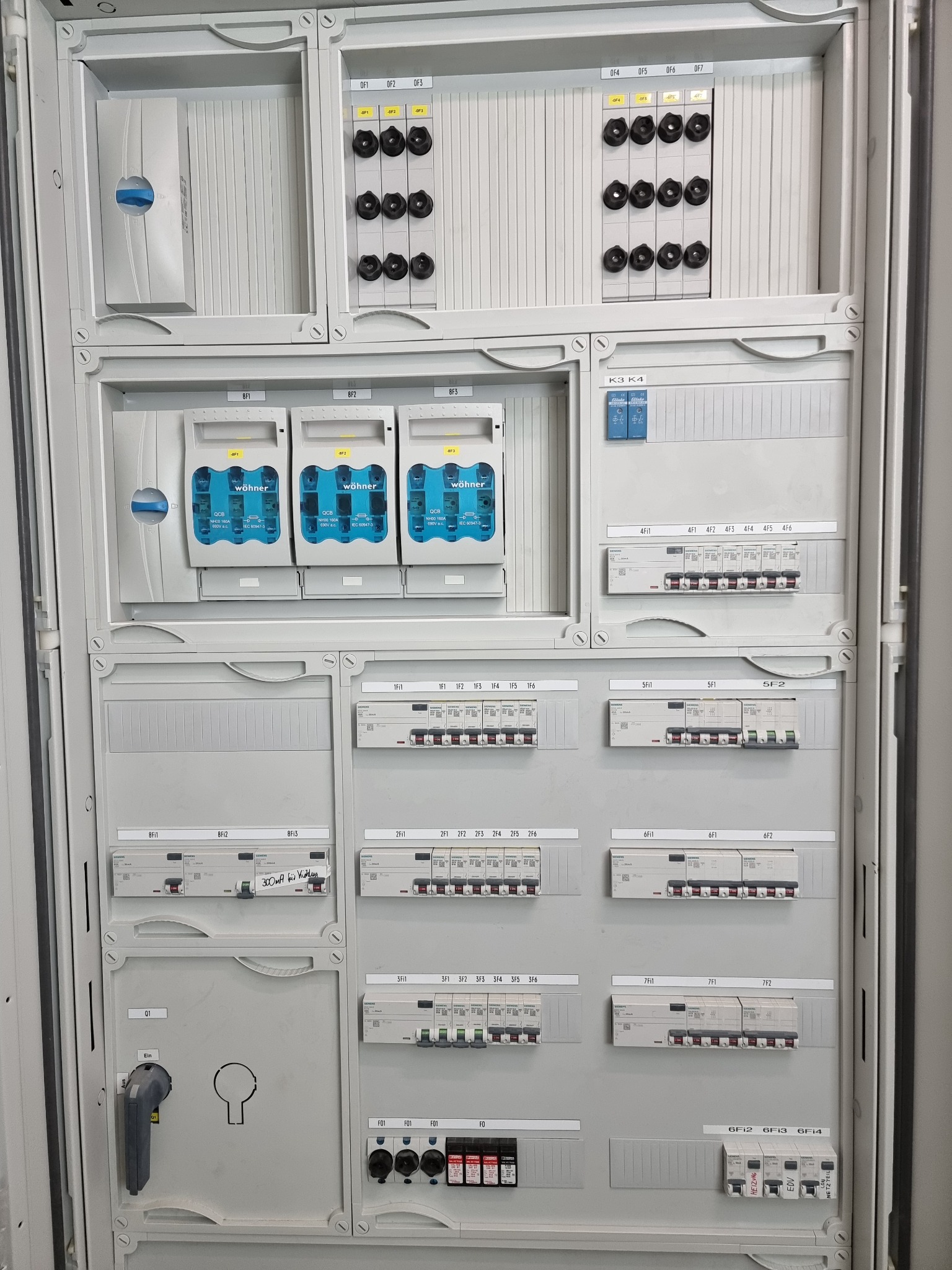 Szara, rozbudowana rozdzielnica elektryczna z bezpiecznikami, wyłącznikami, miernikami i oznaczeniami, widok z przodu, oświetlenie studyjne.