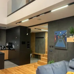 Dwupoziomowe wnętrze z drewnianą podłogą i schodami, czarną zabudową kuchenną, szarą ścianą z obrazem i szarą sofą z żółtą poduszką.