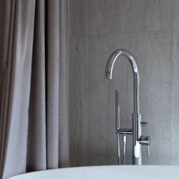 OYAA - Nowoczesna, wolnostojąca wanna z chromowaną armaturą i zasłoną w odcieniach szarości na tle płytek imitujących kamień.