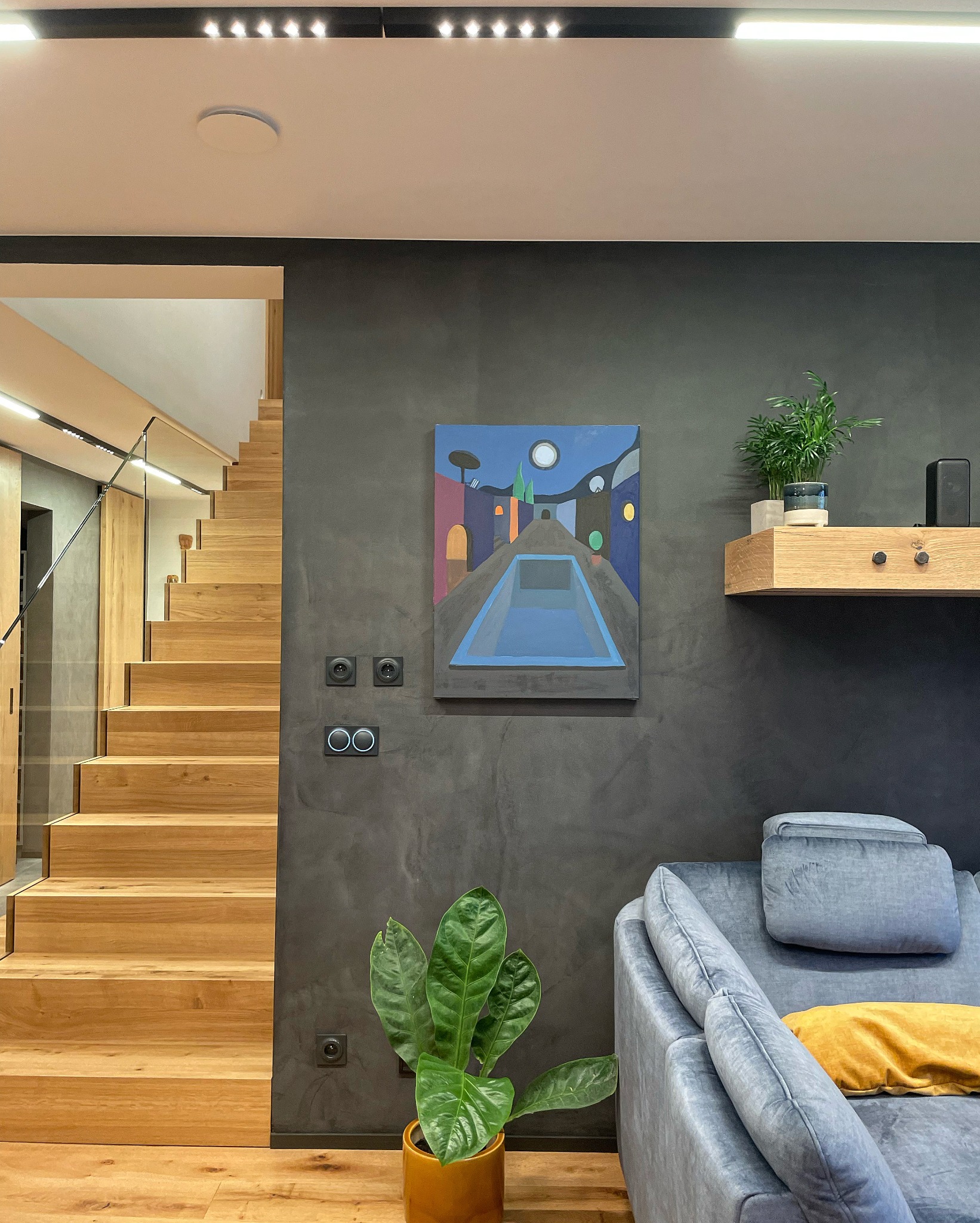 Nowoczesny salon z drewnianymi schodami, szarą ścianą z abstrakcyjnym obrazem, roślinami doniczkowymi i szarą sofą z żółtą poduszką.