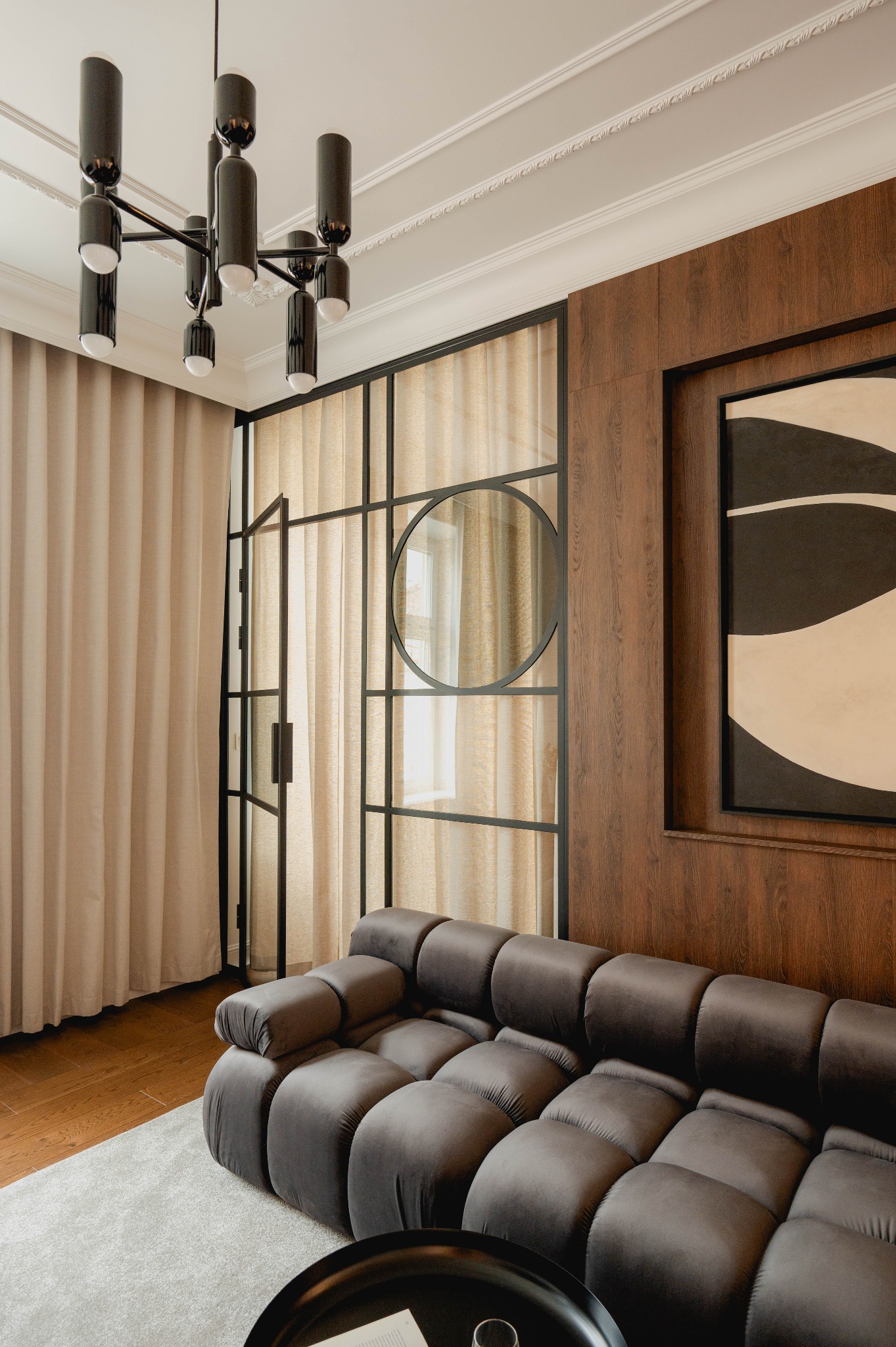 Salon w stylu nowoczesnym z modułową sofą w kolorze grafitowym, drewnianą boazerią, czarną lampą sufitową i przeszkloną ścianą z okrągłym elementem dekoracyjnym.