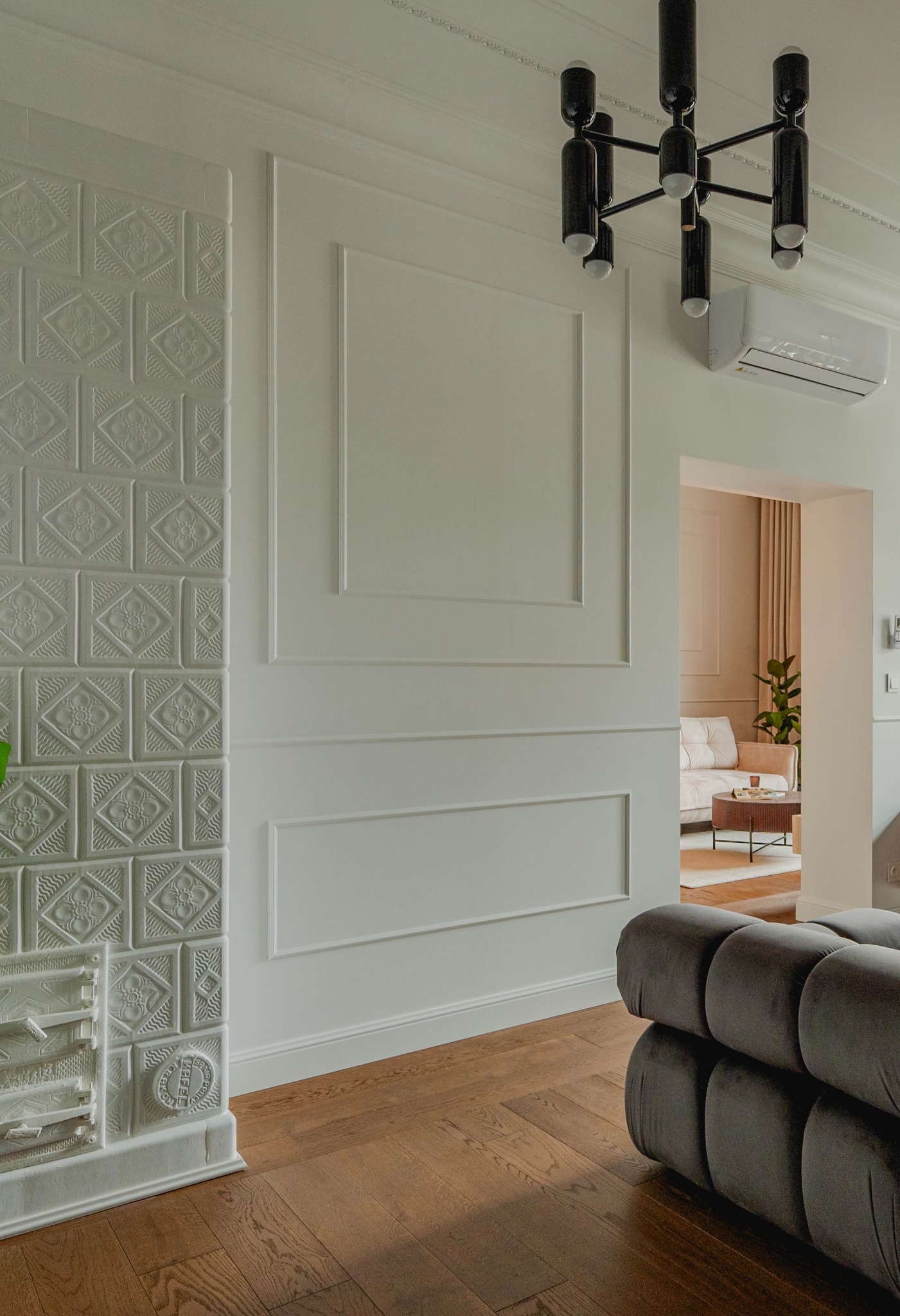 Jasne wnętrze z dekoracyjną ścianą z ceramicznych kafli, panelami ściennymi, nowoczesnym żyrandolem i fragmentem szarej sofy, widok na salon w głębi.