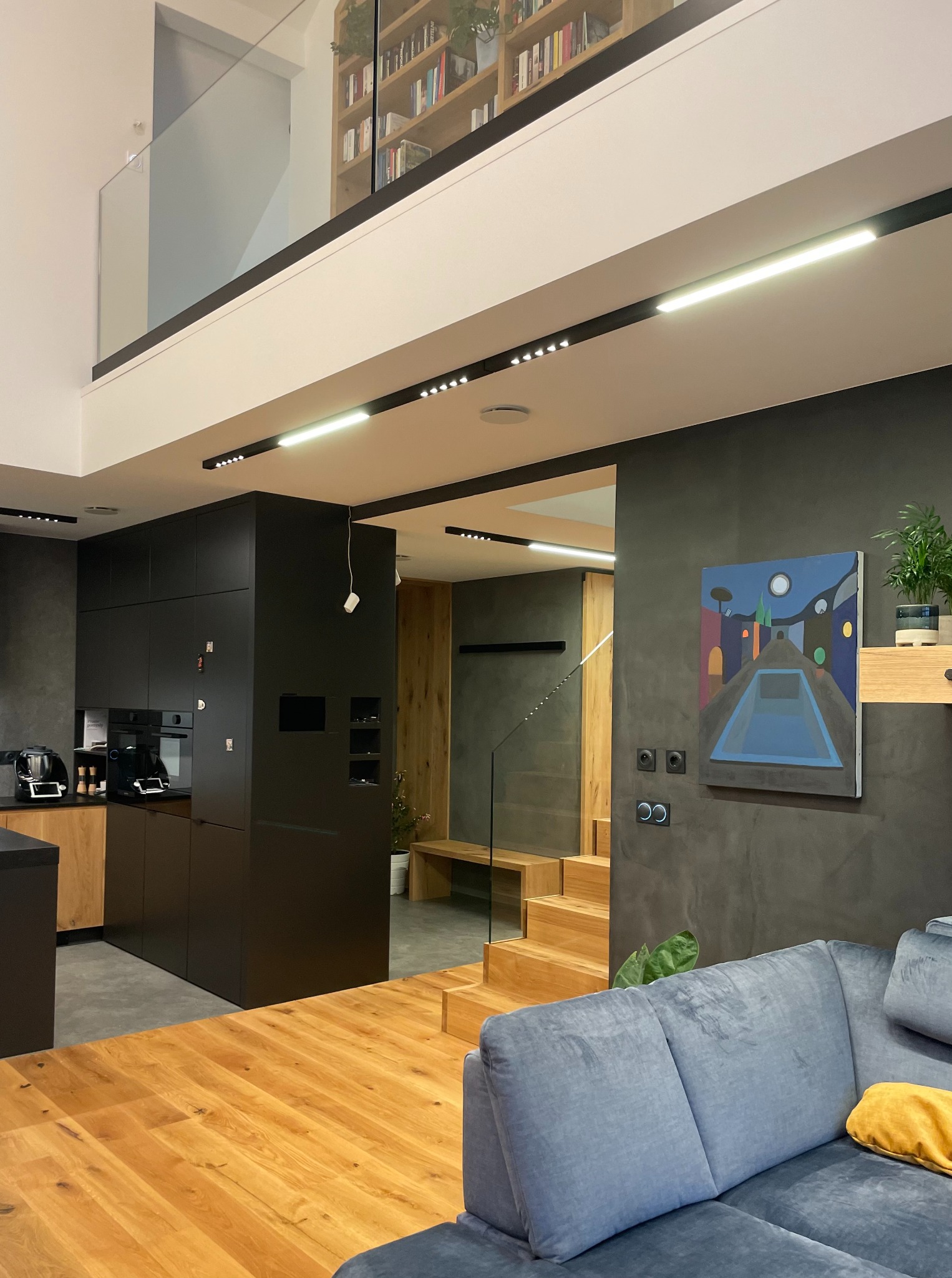 Dwupoziomowe wnętrze z drewnianą podłogą i schodami, czarną zabudową kuchenną, szarą ścianą z obrazem i szarą sofą z żółtą poduszką.