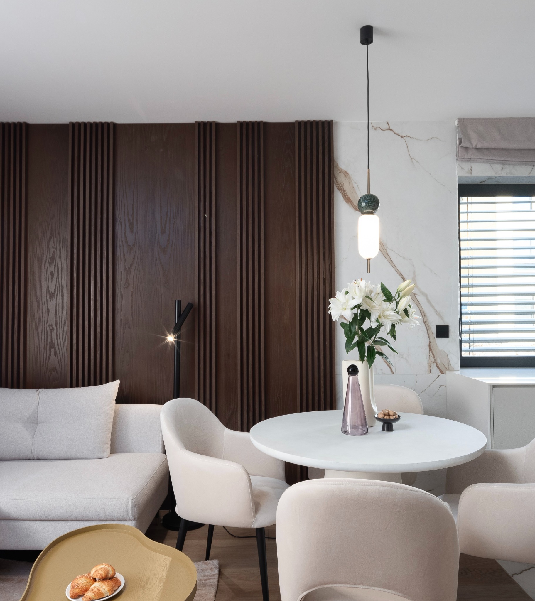Jasne wnętrze salonu z jadalnią: kremowa sofa, okrągły biały stół otoczony beżowymi krzesłami, dekoracyjna ciemnobrązowa ściana z pionowych listew oraz marmurowa ściana z delikatnym wzorem.