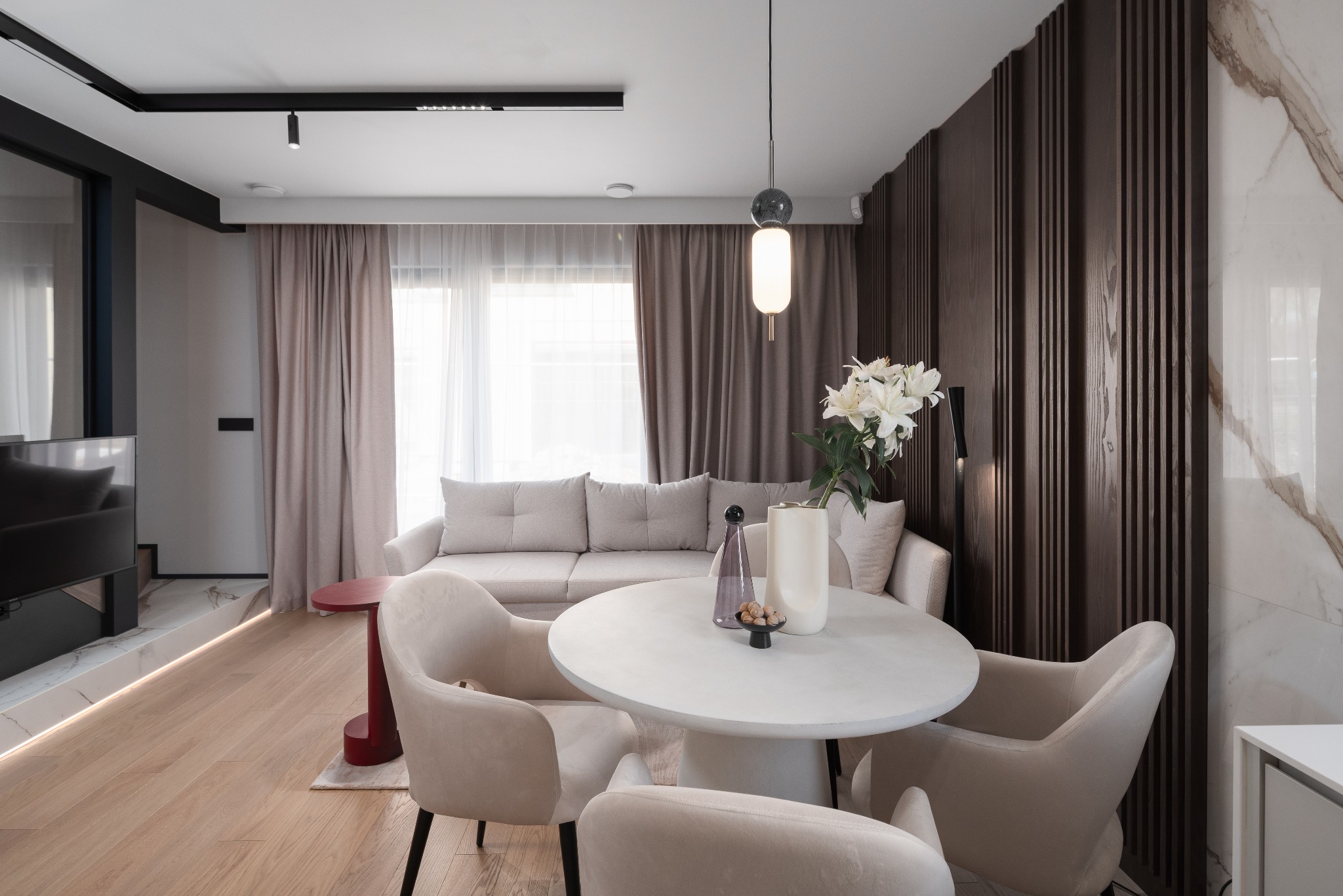 Jasne wnętrze salonu z okrągłym stołem, beżowymi krzesłami, sofą i dekoracyjną ścianą z drewnianych paneli, oświetlone wiszącą lampą i światłem dziennym.