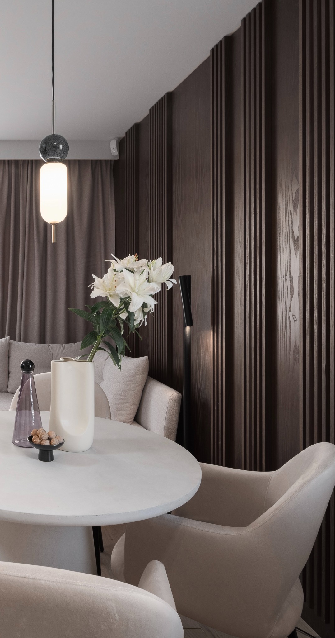 Elegancka jadalnia z okrągłym stołem z betonu architektonicznego, beżowymi krzesłami, dekoracyjną ścianą z drewnianych paneli, białymi liliami w ceramicznym wazonie i nowoczesną lampą wiszącą...