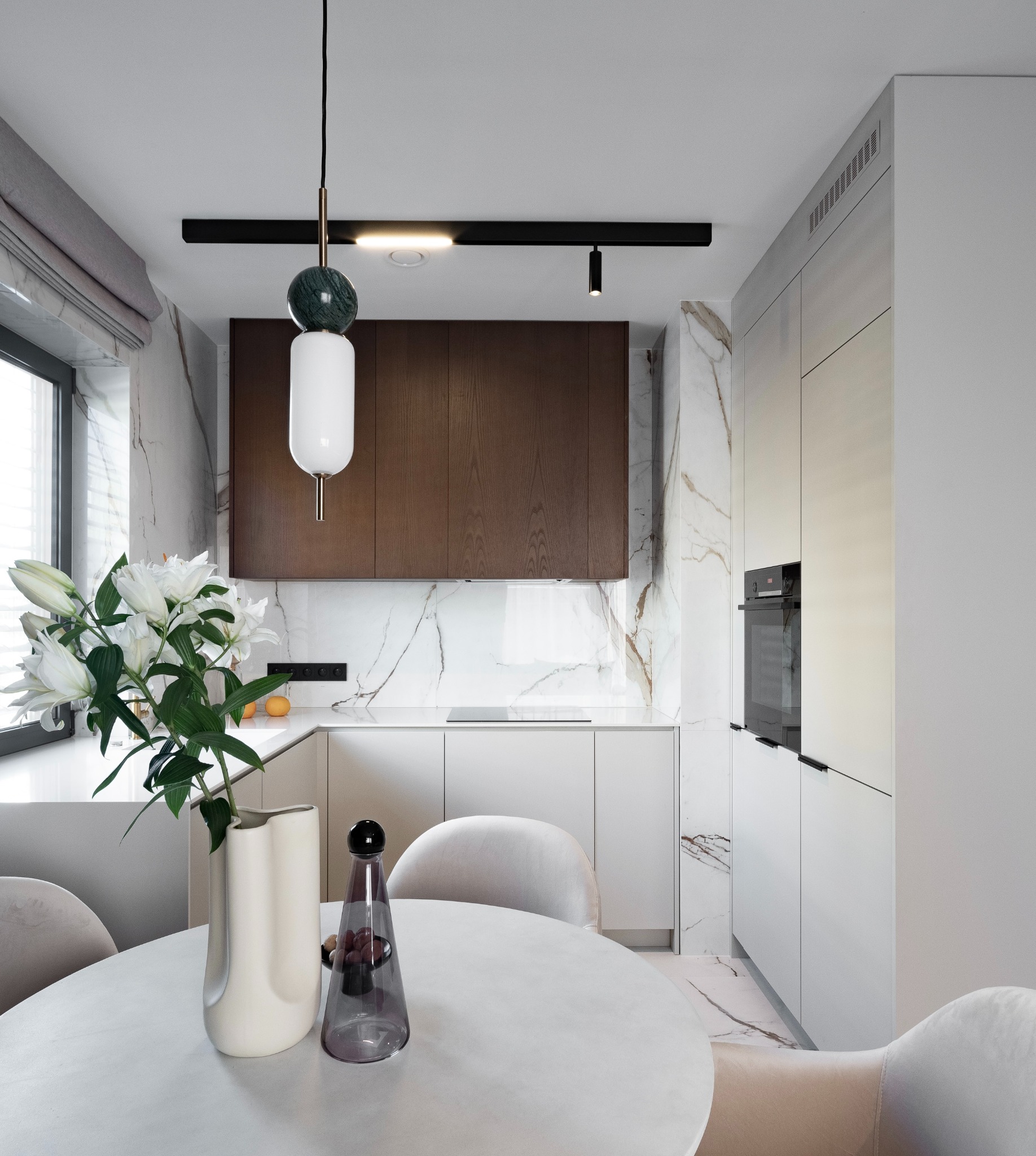 Nowoczesna, minimalistyczna kuchnia z marmurowym akcentem, okrągłym stołem z bukietem białych lilii i designerską lampą wiszącą z elementami marmuru.