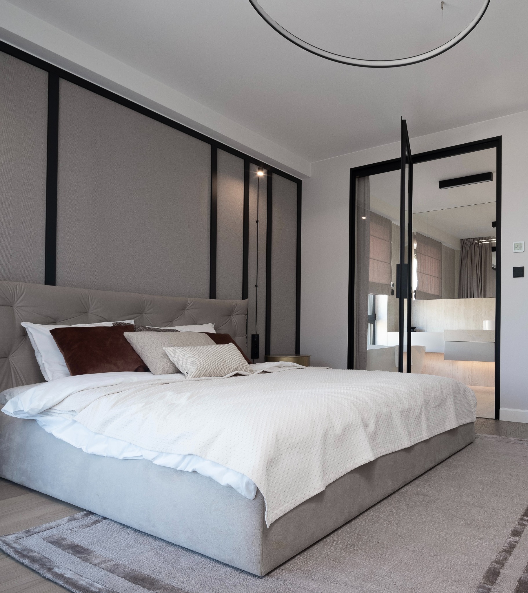 Elegancka sypialnia z łazienką en-suite oddzieloną szklanymi drzwiami, szara tapicerowana ściana za wezgłowiem łóżka z czarnymi pionowymi akcentami, minimalistyczna lampa sufitowa w kształcie okręgu.