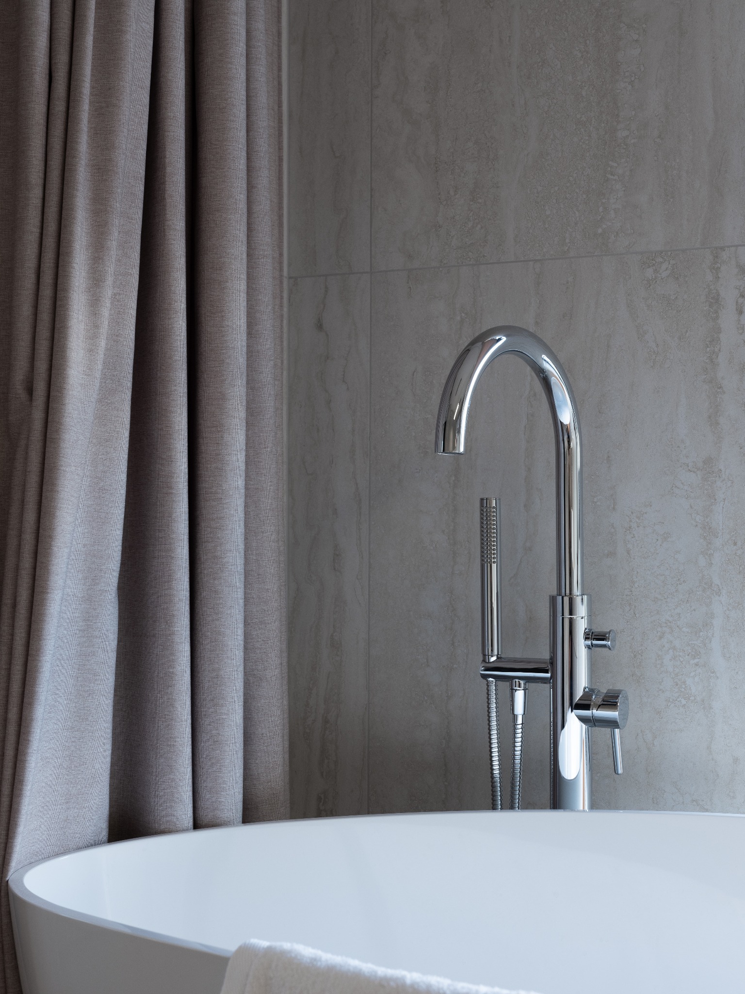 Nowoczesna, wolnostojąca wanna z chromowaną armaturą i zasłoną w odcieniach szarości na tle płytek imitujących kamień.